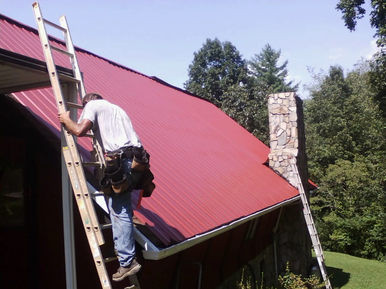 Roofer on a ladder beside a red metal roof and stone chimney — Soffit & Fascia Repair by Roofing Brothers in Weyauwega